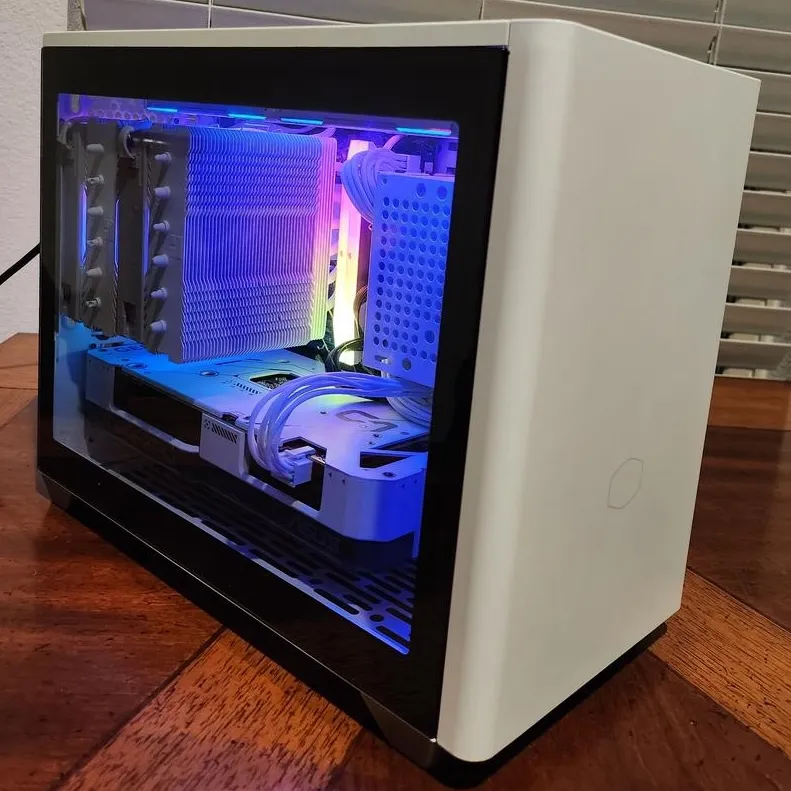 White Cooler Master NR200P mini-ITX case on a wooden table, its glass panel showing a white dual-tower Thermalright air cooler, white cables, and blue RGB lighting.