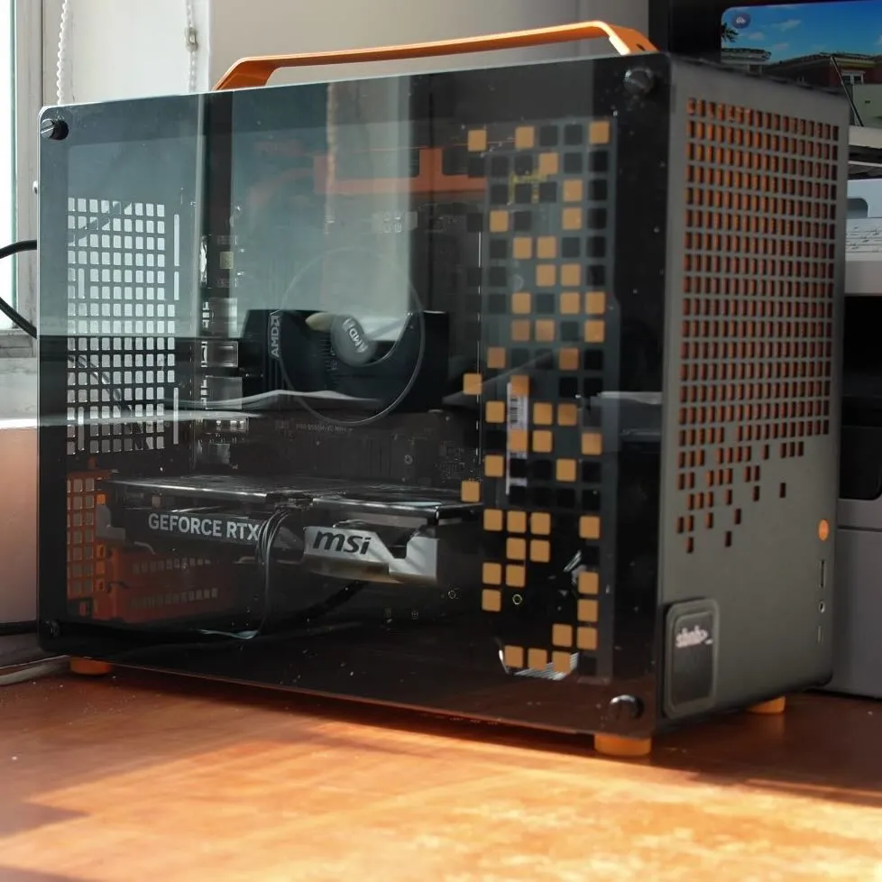 Black JONSBO Z20 case with an orange carry handle and orange-accented perforated panel, its glass side showing an MSI GeForce RTX graphics card and an AMD stock cooler, on a sunlit wooden desk.